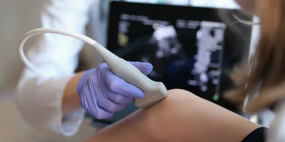 Lekarz lub diagnosta przeprowadza badanie stawu kolanowego przy pomocy urządzenia do USG. Widoczna jest sonda przystawiona do kolana, która umożliwia przekazanie obrazu i sprawdzenie stanu stawu kolanowego.